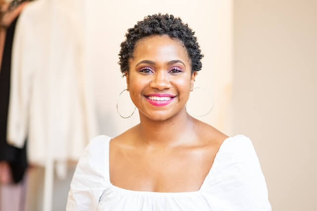 Photo of a smiling woman with short black curly hair and a white peasant style top standing in a clothing store.