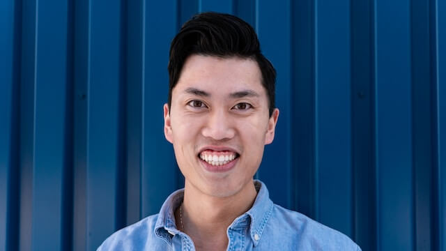 Close up photo of a smiling man with dark hair on a blue backdrop.